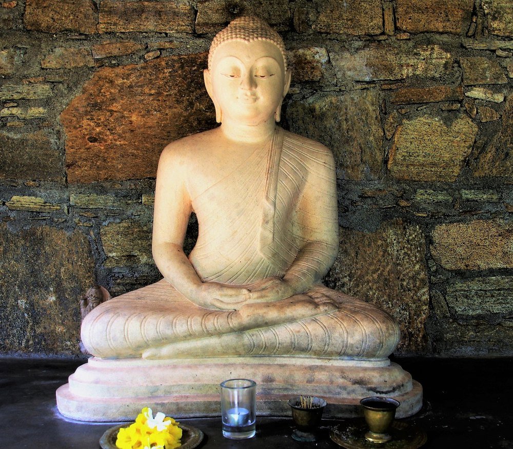 A seated stone Buddha with a lit candle, incense and a dish of yellow flowers